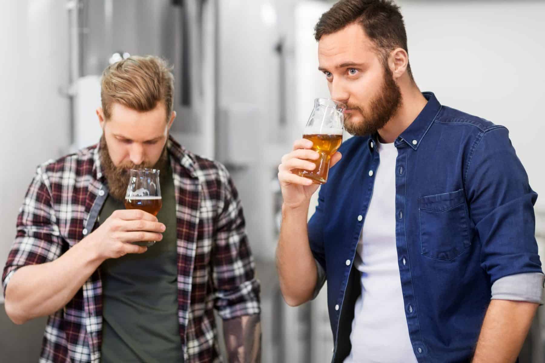 Two men with beards are standing indoors, each holding and smelling a glass of beer, appearing to evaluate its aroma.